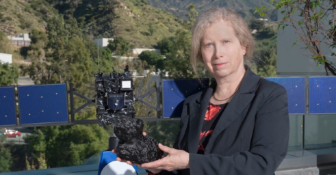 An older white woman in a black blazer standing in front of a model of a spaceship and holding a model of an outer solar system object.